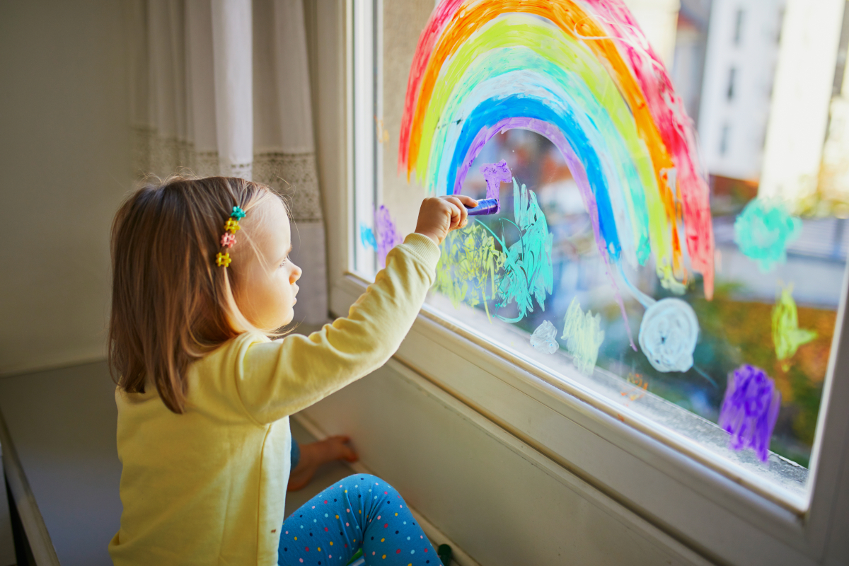 Adorable toddler girl drawing rainbow on window glass